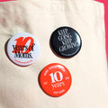 Three buttons on a tote bag against a hot pink background