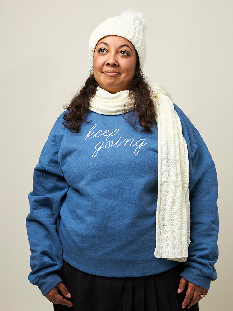 Front view of woman wearing a light blue crewneck sweatshirt with the phrase Keep Going embroidered in white in a cursive font across the chest.