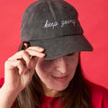 Front view of woman wearing a gray ballcap with white embroidery that reads KEEP GOING in script font.
