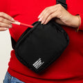 Woman pulling pen out of an all black nylon belt bag with an adjustable strap, zip closure, and small white Moms Demand Action Logo printed on the bottom left corner.