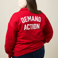 Back view of woman wearing a red quarter-zip collared sweatshirt with the phrase DEMAND ACTION printed in large white all caps text across the back.