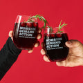 Two hands holding up clear stemless drinking glasses with a Moms Demand Action logo etched into the surface of each, filled with dark red juice and cranberries and garnished with rosemary.