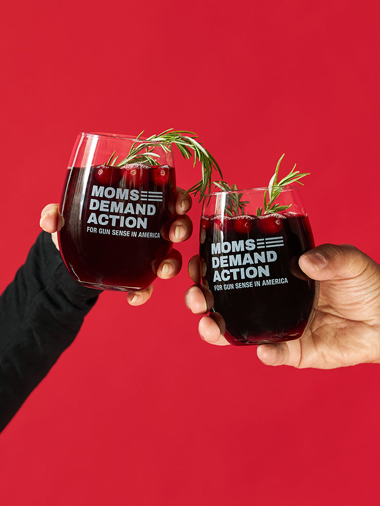 Two hands holding up clear stemless drinking glasses with a Moms Demand Action logo etched into the surface of each, filled with dark red juice and cranberries and garnished with rosemary.