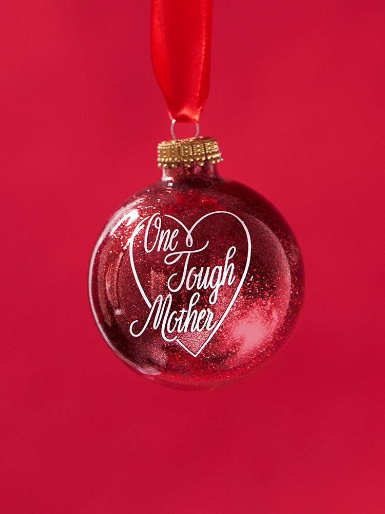 Red, glittery, round plastic ornament with One Tough Mother printed in script font in white on one side.