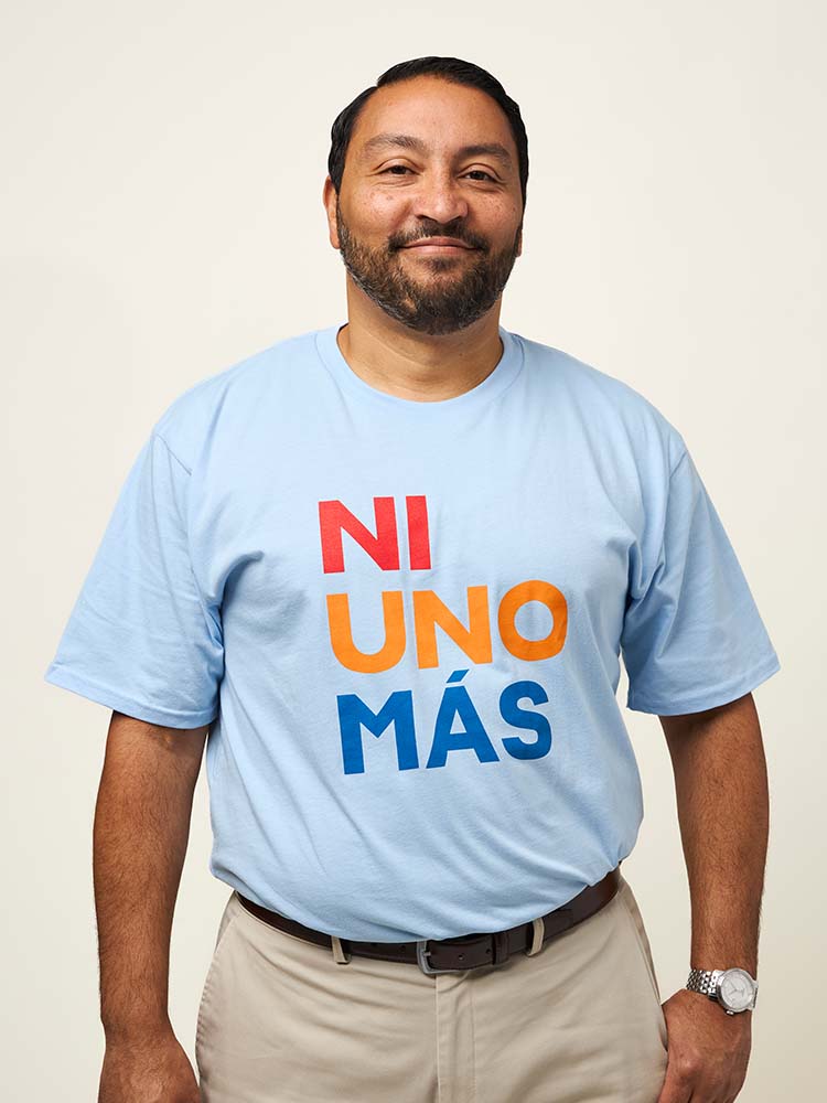 Front view of a man wearing a light blue short sleeve unisex t-shirt with print that reads NI UNO MÁS in large, uppercase, red, orange, and blue letters.
