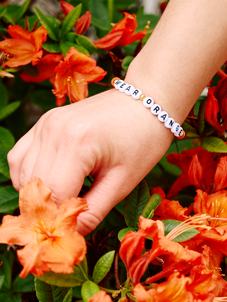 A hand wearing a beaded friendship bracelet that reads "WEAR ORANGE" picks a flower.