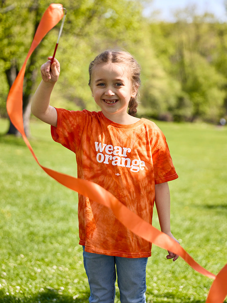A girl wearing an orange tie-dye short-sleeve unisex kids t-shirt with a white Wear Orange logo printed across the chest smiles as she twirls an orange ribbon in a grassy field.